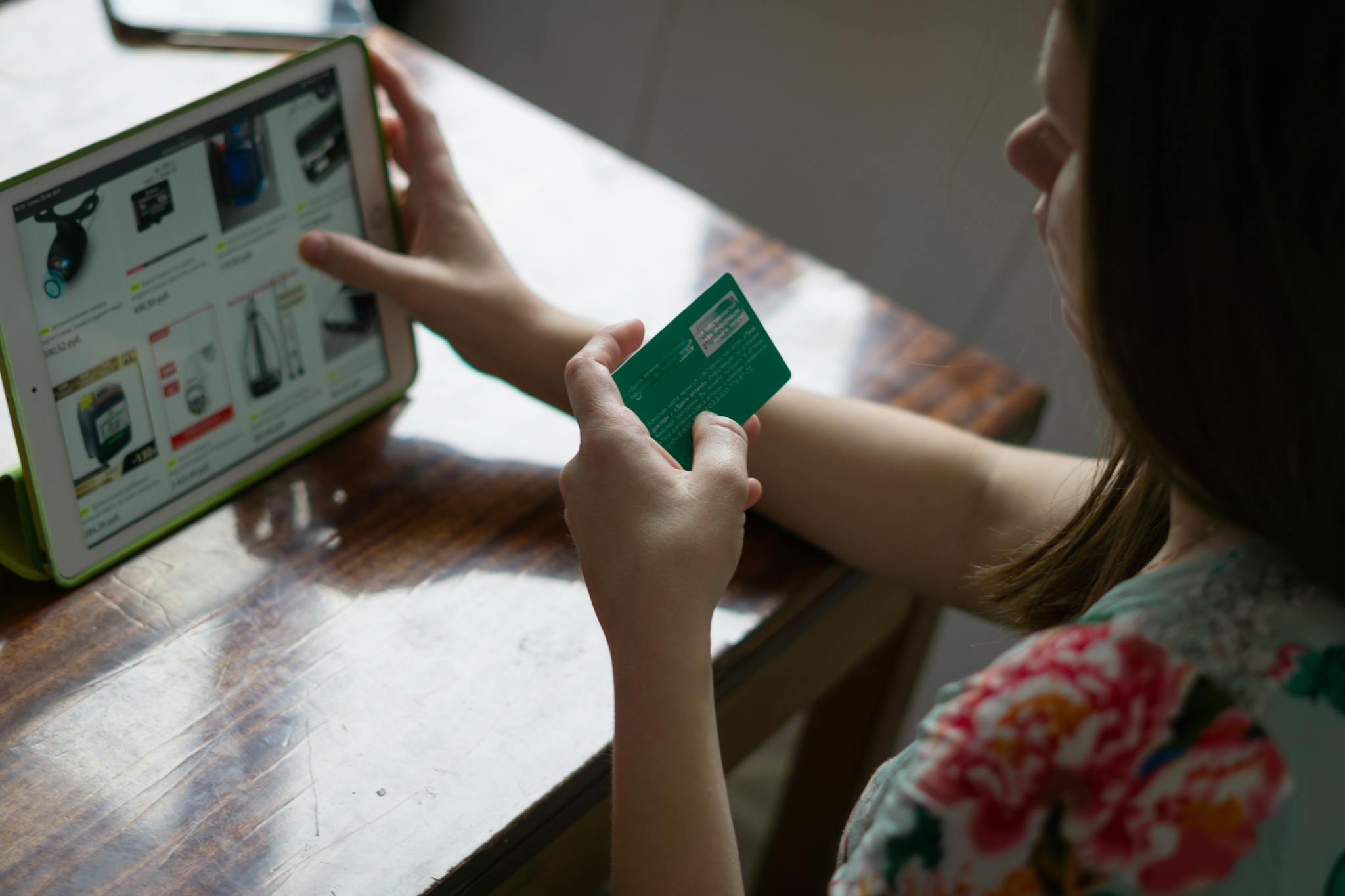 A woman uses a credit card for online shopping on a tablet at a wooden table.